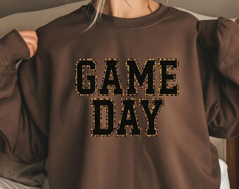 Può includere: Una felpa marrone con la scritta "GAME DAY" in grandi lettere nere con un motivo leopardato. La felpa ha maniche lunghe e girocollo.