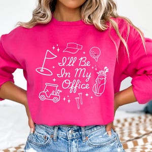 Op de afbeelding: Een felroze sweatshirt met de tekst "I'll Be In My Office" en witte golfafbeeldingen. De afbeeldingen omvatten een golflag, vizier, golfbal, golfkar, golftas en tees.