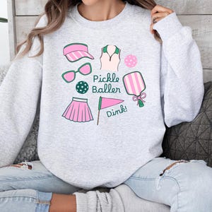 May include: A gray sweatshirt with a pink and green pickleball design. The design includes a pickleball paddle, a ball, a visor, sunglasses, a skirt, and a shirt. The text "Pickle Baller" and "Dink!" are also included in the design.