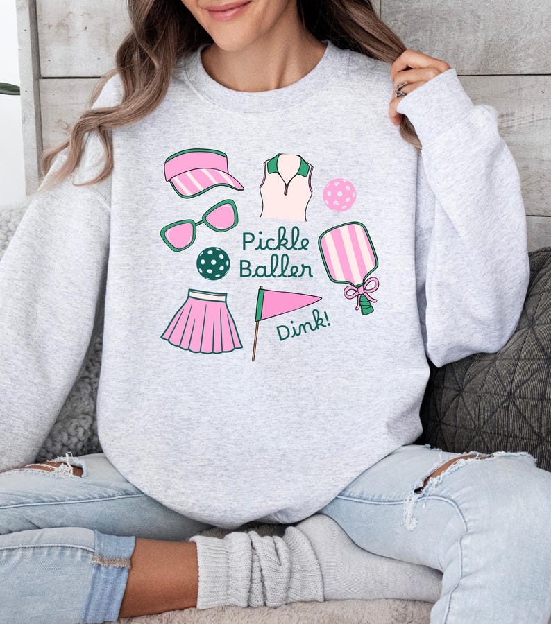 May include: A gray sweatshirt with a pink and green pickleball design. The design includes a pickleball paddle, a ball, a visor, sunglasses, a skirt, and a shirt. The text "Pickle Baller" and "Dink!" are also included in the design.