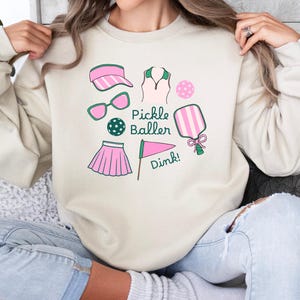 May include: A cream-colored sweatshirt with a pink and green pickleball design. The design includes a pickleball paddle, a ball, a visor, sunglasses, a skirt, a shirt, and a flag with the text "Pickle Baller" and "Dink!"