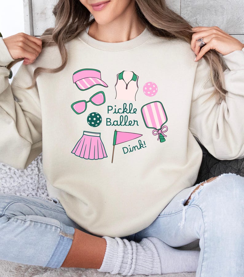May include: A cream-colored sweatshirt with a pink and green pickleball design. The design includes a pickleball paddle, a ball, a visor, sunglasses, a skirt, a shirt, and a flag with the text "Pickle Baller" and "Dink!"