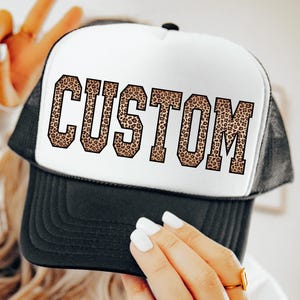 May include: A black and white trucker hat featuring the word "CUSTOM" in a leopard print pattern. The hat has a black peak and mesh back, with a white front panel. The text is in a bold, block letter style.