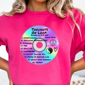 May include: A bright pink sweatshirt featuring a graphic of a CD with the text "THOUGHTS ON LOOP" and a list of thoughts. The CD design includes a peace sign, yin-yang symbol, and a heart. The sweatshirt is a vibrant, solid color.
