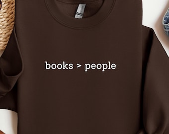 May include: A dark brown sweatshirt with the text "books > people" in white. The sweatshirt is a crew neck style, with a ribbed collar and cuffs. The Gildan brand tag is visible.