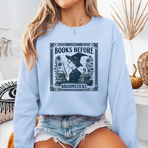 May include: Light blue sweatshirt with a graphic featuring a witch with a hat, holding a wand, and surrounded by books and flowers. The text reads "BOOKS BEFORE BROOMSTICKS" in a decorative frame.