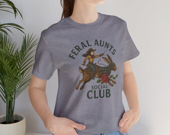 May include: A heather gray t-shirt with the words "FERAL AUNTS SOCIAL CLUB" in a circular design. The graphic features a cowgirl riding a rabbit with a rose. The shirt is made of a soft material.