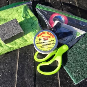 May include: A green cloth, a black sponge, a small tin of Mind Your Blade's Wax, a pair of green scissors, a green scrubby sponge, and a clear plastic bag with a red and blue tape inside.