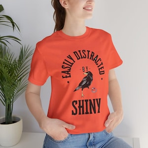 Puede incluir: Camiseta naranja con un gráfico de cuervo negro y el texto "Easily Distracted by Shiny".