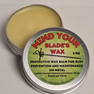 Könnte beinhalten: Eine silberne Dose mit einem grün-roten Etikett, auf dem "Mind Your Blade's Wax 1 oz" steht. Das Etikett trägt außerdem den Text "Protective Wax Balm for Rust Prevention and Maintenance on Metal. Locally sourced beeswax and natural oils by Blackram Farm. Find at: Findingjoysinlife.etsy.com".