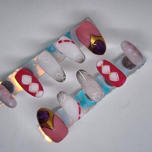 May include: A set of ten false nails with different designs. The nails are arranged on a blue and white display card. The designs include white flowers, red and white squares, and a purple and gold design.