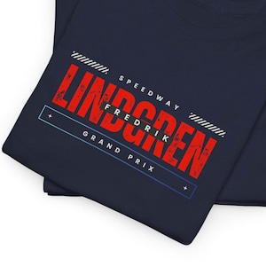 Può includere: T-shirt blu navy con un design grafico rosso e bianco. Il design include il nome "LINDGREN" a grandi lettere rosse, con "SPEEDWAY" e "GRAND PRIX" in testo bianco. È incluso anche il testo "FREDRIK".