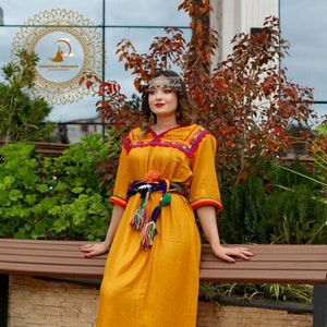 May include: A woman wearing a long, mustard yellow dress with colorful embroidery and a beaded belt. She is wearing a silver headpiece and white sneakers. The dress has a slit on the side and a colorful trim around the bottom.