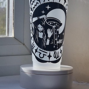 Może przedstawiać: Półprzezroczysty plastikowy kubek z przezroczystą pokrywką i słomką, ozdobiony czarną naklejką winylową ze znakiem zodiaku Wodnik. Wzór przedstawia postać wylewającą wodę z naczynia na tle dużego księżyca, otoczoną różnymi symbolami zodiaku.