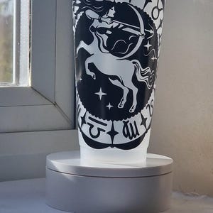 Może przedstawiać: Półprzezroczysty plastikowy kubek na zimne napoje z pokrywką i słomką, ozdobiony czarnym wzorem znaku zodiaku Strzelec przedstawionym jako centaur-łucznik, otoczony kolistą obwódką z różnymi symbolami astrologicznymi i małymi gwiazdkami.