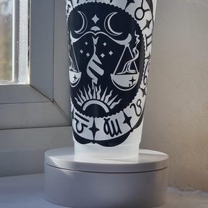 Może przedstawiać: Półprzezroczysty plastikowy kubek na zimne napoje z przezroczystą pokrywką i słomką, ozdobiony czarną naklejką. Wzór przedstawia symbol zodiakalnej Wagi otoczony okrągłą obwódką z różnymi znakami zodiaku oraz motywami gwiazd.