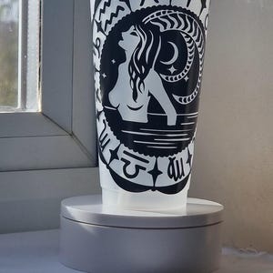 Może przedstawiać: Półprzezroczysty plastikowy kubek z przezroczystą pokrywką i słomką, ozdobiony czarnym wzorem graficznym przedstawiającym postać wyłaniającą się z wody, otoczoną symbolami zodiaku, w tym znakami Ryb i Barana w okrągłym układzie.