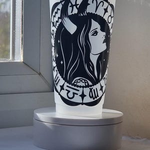 Może przedstawiać: Półprzezroczysty biały plastikowy kubek na zimne napoje z przezroczystą pokrywką i słomką, ozdobiony czarnym wzorem graficznym przedstawiającym rogatą postać z profilu, otoczoną okrągłą ramką z symbolami zodiaku i małymi gwiazdkami.