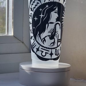 Może przedstawiać: Półprzezroczysty plastikowy kubek na zimne napoje z pokrywką i słomką, ozdobiony czarnym wzorem graficznym przedstawiającym postać trzymającą skorpiona, otoczoną różnymi symbolami zodiaku ułożonymi w kolisty wzór wokół centralnej postaci.
