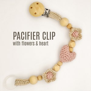 May include: A beige crocheted pacifier clip with wooden beads, a pink crocheted heart, and two pink crocheted flowers. The clip is attached to a wooden ring. The text "PACIFIER CLIP with flowers & heart" is printed below the clip.