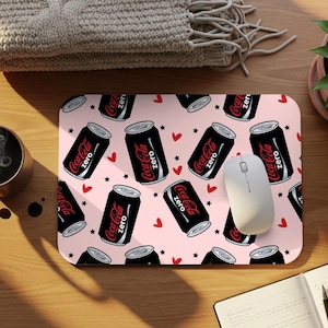 May include: A rectangular mousepad with a pink background featuring a repeating pattern of black Coca-Cola Zero cans, red hearts, and black stars. A white computer mouse rests on the pad.