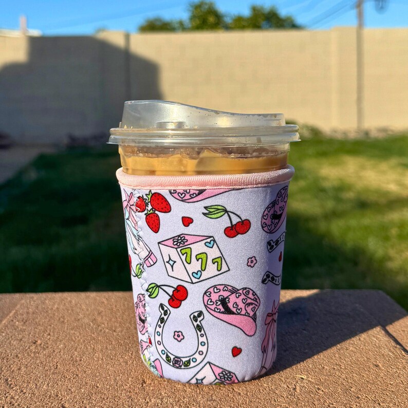May include: A pink and white insulated cup sleeve with a pattern of strawberries, horseshoes, dice, and cowboy hats. The sleeve is on a brown surface.