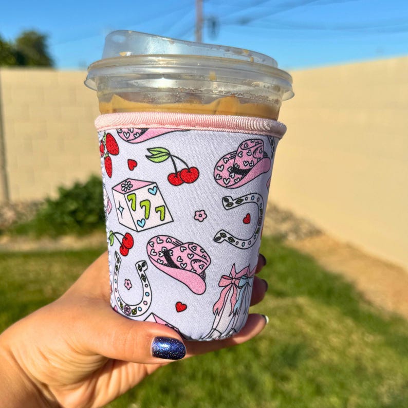 May include: A pink and white reusable coffee cup sleeve with a pattern of dice, horseshoes, cherries, and cowboy hats. The sleeve is made of neoprene and has a zipper closure.