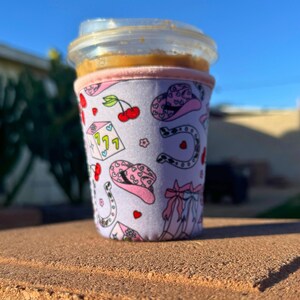 May include: A pink and white insulated cup sleeve with a pattern of cowboy hats, horseshoes, cherries, and dice. The sleeve is on a brown surface.