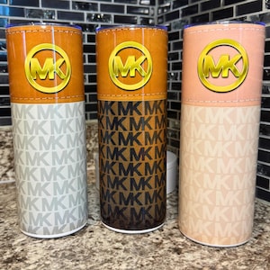May include: Three stainless steel tumblers with the "MK" logo. The top sections are brown, with a gold logo. The middle and bottom sections feature the "MK" logo in different patterns and colors: white, brown, and peach.