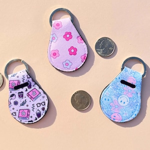 May include: Three teardrop-shaped keychains with silver key rings. One keychain is light purple with pink flowers, another is light blue with smiley faces and flowers, and the third is lavender with books and coffee cups.