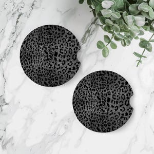 May include: Two round car coasters with a black and silver leopard print design. Each coaster has a small notch for easy removal from a car cup holder. The coasters are made of a durable material.