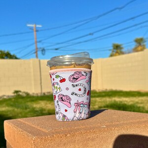 May include: A pink and white coffee sleeve with a pattern of cowboy hats, horseshoes, cherries, and dice. The sleeve is on a clear plastic cup.