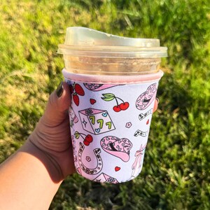 May include: A pink and white neoprene cup sleeve with a design of cherries, dice, horseshoes, and cowboy hats. The sleeve is on a clear plastic cup with a brown liquid inside.