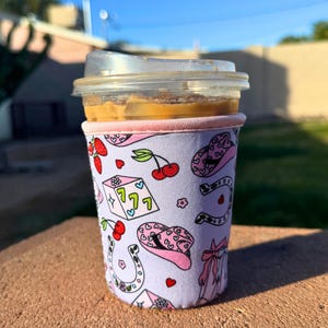 May include: A pink and white insulated cup sleeve with a pattern of cherries, dice, horseshoes, and cowboy hats. The sleeve is on a coffee cup with a brown iced coffee drink.