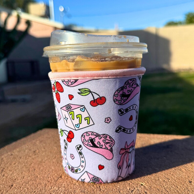 May include: A pink and white insulated cup sleeve with a pattern of cherries, dice, horseshoes, and cowboy hats. The sleeve is on a coffee cup with a brown iced coffee drink.