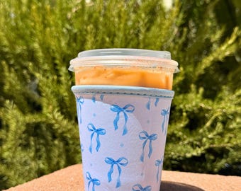 May include: A clear plastic cup filled with an orange beverage, topped with a clear lid, and encased in a white cup sleeve. The sleeve is decorated with blue bows and small blue dots. The background is a blurred green bush.