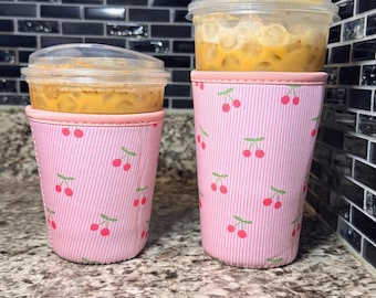 May include: Two pink and white striped insulated cup sleeves with a cherry pattern. The sleeves are on a countertop with a light brown speckled pattern.