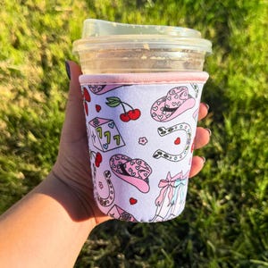 May include: A pink and white neoprene coffee cup sleeve with a pattern of cherries, horseshoes, dice, and cowboy hats. The text "777" is on the dice.