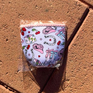 May include: A pink and white neoprene can cooler with a pattern of pink cowboy hats, horseshoes, cherries, and dice. The cooler has a pink zipper and a white background.
