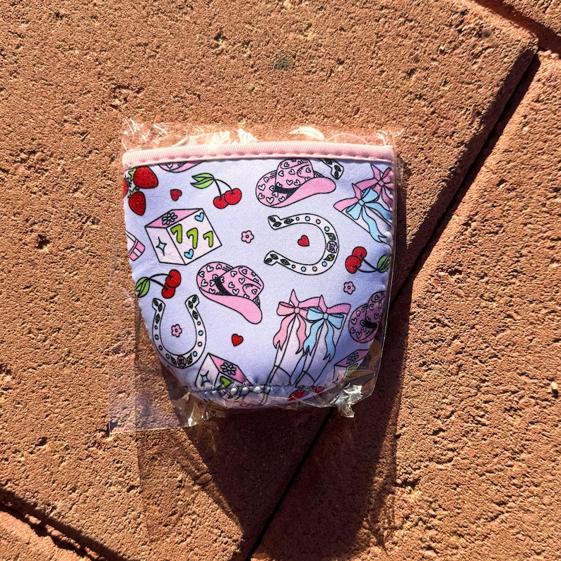 May include: A pink and white neoprene can cooler with a pattern of pink cowboy hats, horseshoes, cherries, and dice. The cooler has a pink zipper and a white background.