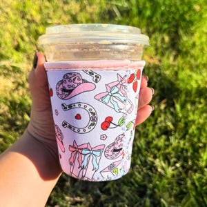 May include: A pink and white insulated cup sleeve with a pattern of cowboy hats, horseshoes, cherries, and bows. The sleeve is designed to fit a standard-sized cup.