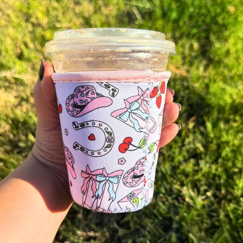 May include: A pink and white insulated cup sleeve with a pattern of cowboy hats, horseshoes, cherries, and bows. The sleeve is designed to fit a standard-sized cup.