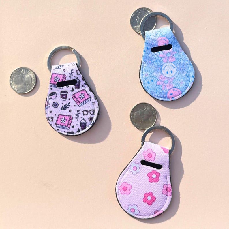 May include: Three keychains with silver key rings. One keychain is lavender with a pattern of books, coffee cups, and glasses. Another is light blue with a floral and smiley face pattern. The third is pink with a flower pattern.