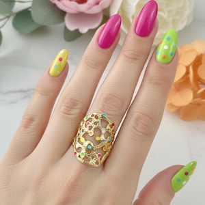 Puede incluir: Un anillo de tono dorado con un diseño intrincado de celosía calada. La estructura metálica está adornada con pequeños puntos de esmalte de colores en tonos azul, naranja, verde y rosa, creando un patrón vibrante de lunares multicolor.