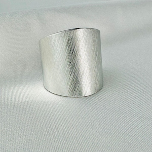 Puede incluir: Un anillo ancho de color plateado con un acabado de metal cepillado. El anillo tiene forma cilíndrica con una parte superior plana y una parte inferior redondeada. La superficie del anillo tiene un patrón diagonal texturizado.
