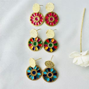 May include: Three pairs of gold-coloured earrings with colourful enamel designs. Each earring features a textured circular stud and a larger, round pendant with a floral pattern in shades of pink, rainbow, and blue. The earrings are displayed on a white surface.