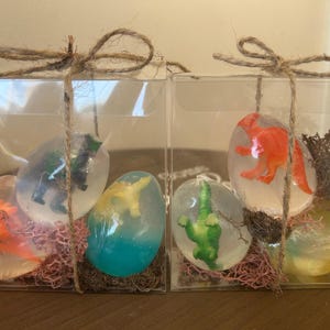 May include: Two clear boxes, each with three dinosaur soaps. The egg-shaped soaps contain a small plastic dinosaur toy. The boxes are tied with twine and presented as a gift.