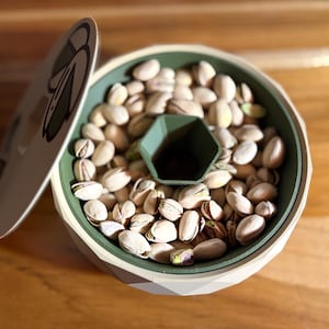May include: A decorative pistachio nut bowl with a lid. The bowl is white and green with a geometric design. The bowl is filled with pistachios and has a green hexagonal compartment in the center.