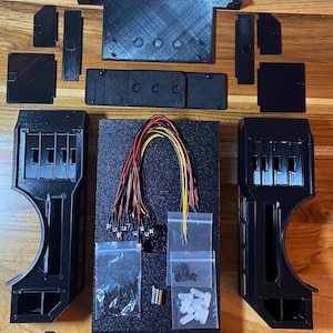 May include: A collection of black 3D-printed components, including panels, brackets, and a central base with wiring and small bags of hardware. The parts are arranged on a wooden surface, suggesting a DIY or electronics project.