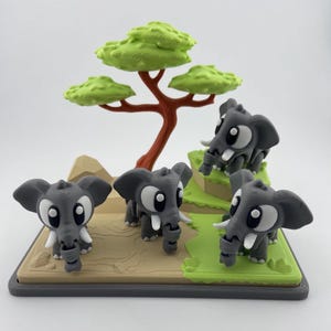 May include: A 3D-printed diorama featuring four gray cartoon elephants with large white eyes and tusks. A green tree and grassy hills are in the background. The base is tan and green.